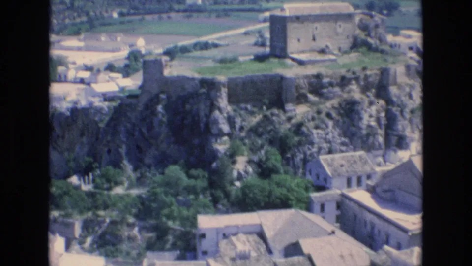 1972 Spain 8mm frame showing stone fortress-like ruins on a rocky outcrop above clustered buildings and distant agricultural fields.