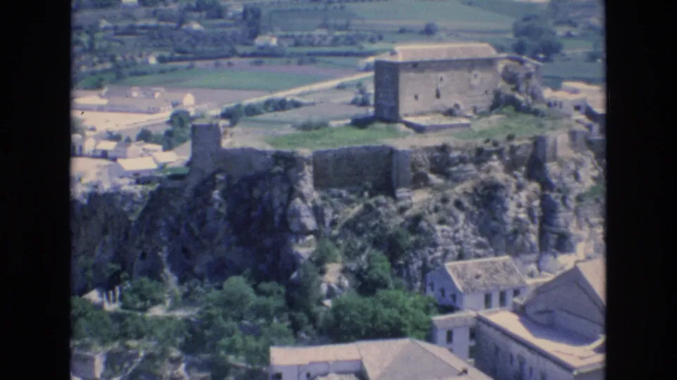 Elevated 1972 Spain home-movie view of rocky hilltop ruins, pale town buildings, trees, and fields beyond the settlement.