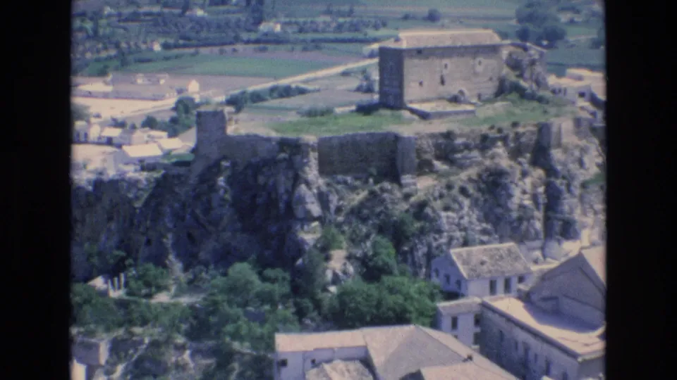 1972 Spain 8mm frame with castle-like stone walls and a rectangular ruin above rooftops, trees, and open fields.