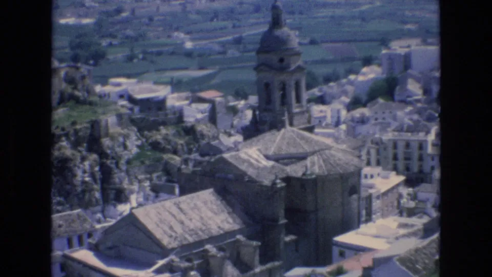 1972 Spain home-movie frame showing a domed church bell tower, tiled roofs, pale buildings, rocky cliffs, and green fields in the background.