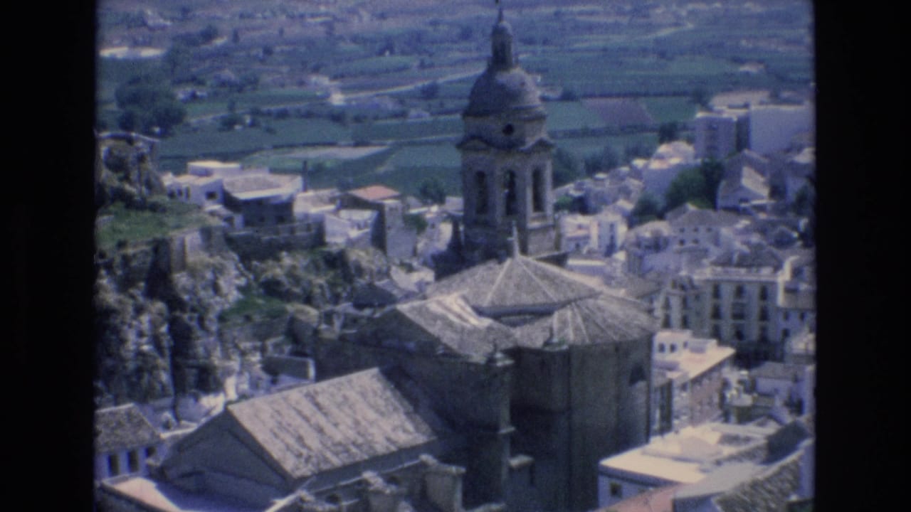 1972 Spain 8mm frame showing an elevated view over a village with a prominent stone church tower and dome, clustered roofs, rocky slopes, and farmland in the distance.