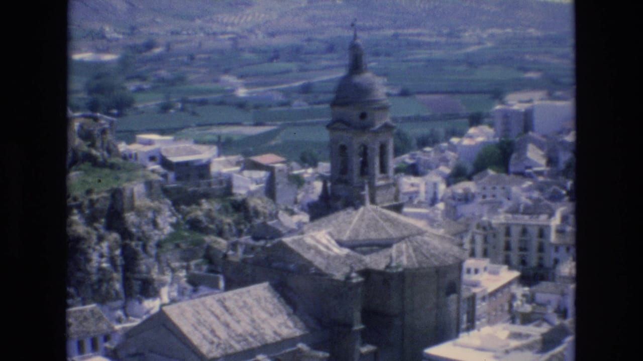 Similar elevated village view with church architecture, rooftops, rocky ground, and surrounding fields.