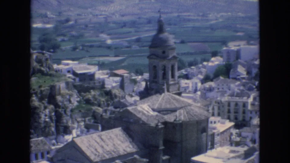 1972 Spain 8mm frame holding on the hillside village, with the church tower near center, pale buildings below, rocky terrain at left, and cultivated fields behind.