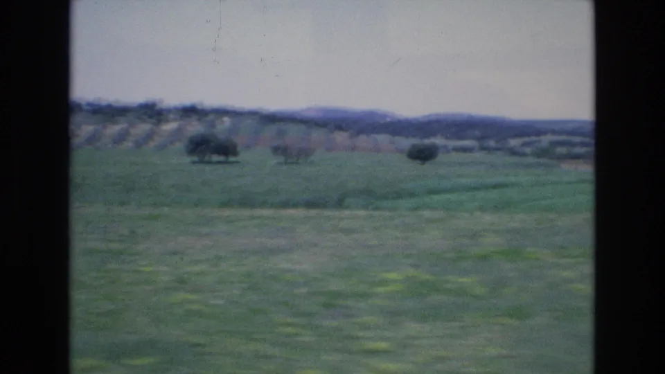 1972 Spain countryside frame showing a broad green field, scattered trees, distant hills, and pale sky with no people visible in this still.
