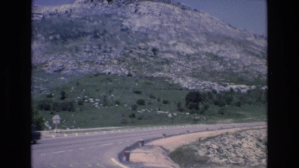 1972 Spain road-trip frame showing a curving paved road, roadside barrier, green slope, and rocky hillside beyond.