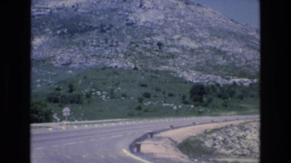1972 Spain 8mm frame showing a road bend with roadside signs, low barriers, green hillside, and exposed rocky terrain.