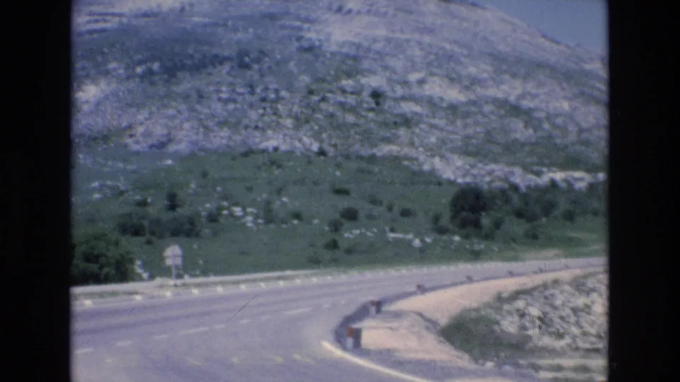 1972 Spain home-movie frame of a curving road, pale shoulder, low roadside barrier, and rocky green hillside under open sky.