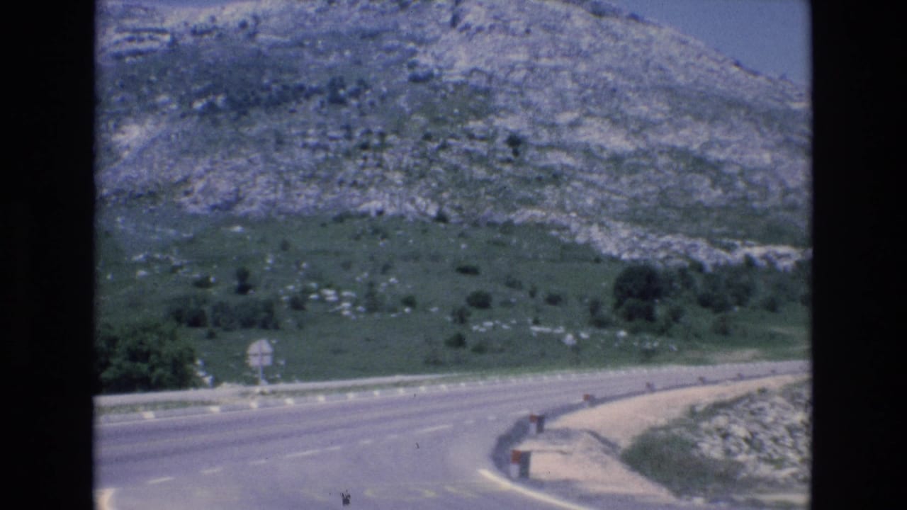 1972 Spain 8mm frame showing a winding paved mountain road, roadside signs, guardrail posts, and rocky green slopes under a bright sky.