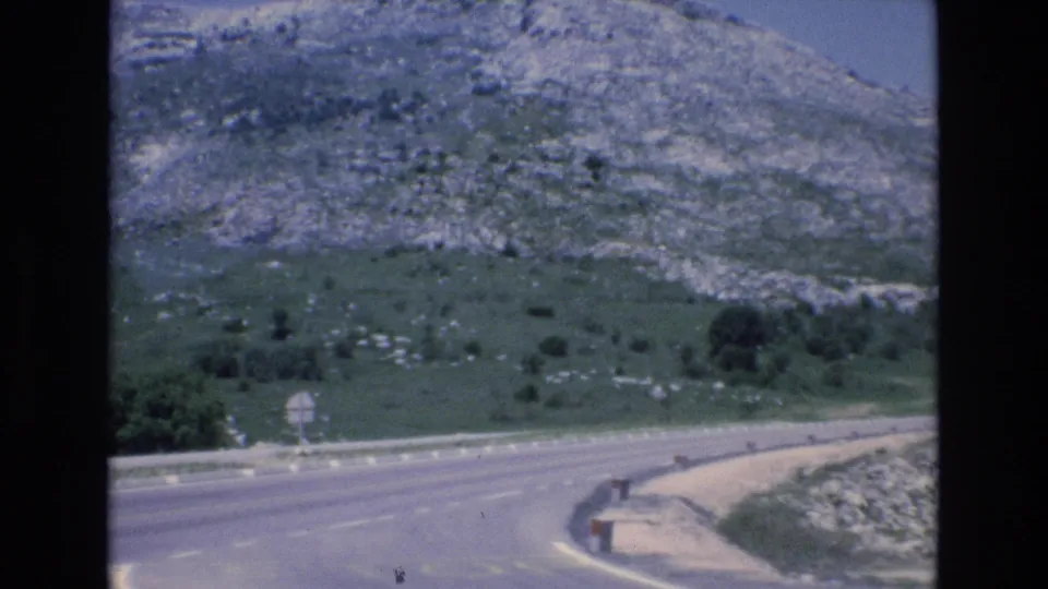 1972 Spain 8mm frame showing a winding paved mountain road, roadside signs, guardrail posts, and rocky green slopes under a bright sky.