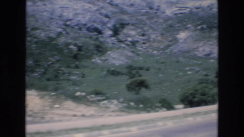 1972 Spain 8mm frame from a moving roadside viewpoint, showing blurred pavement, green hillside, scattered rocks, shrubs, and mountain terrain.