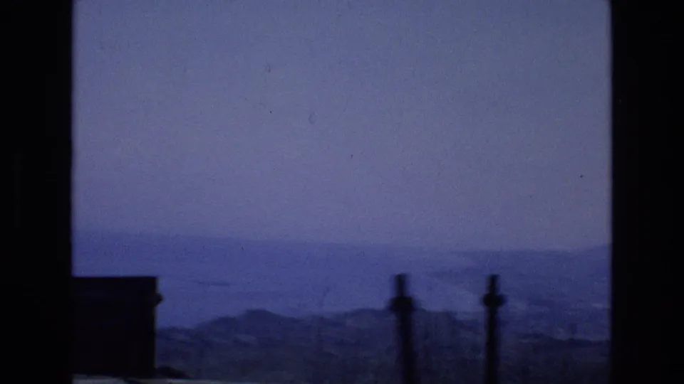 1972 Spain 8mm frame looking from a high roadside or overlook toward the distant sea, hazy sky, coastal hills, and dark foreground posts.