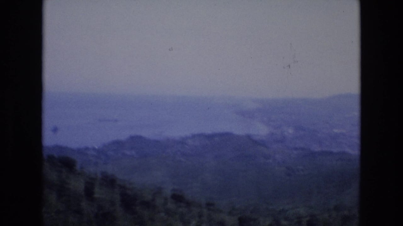 1972 Spain 8mm frame showing a blue-tinted hazy coastal panorama from an elevated viewpoint, with dark hills in the foreground and distant water beyond.