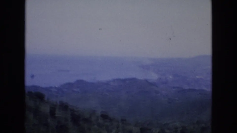 1972 Spain 8mm frame showing a blue-tinted hazy coastal panorama from an elevated viewpoint, with dark hills in the foreground and distant water beyond.
