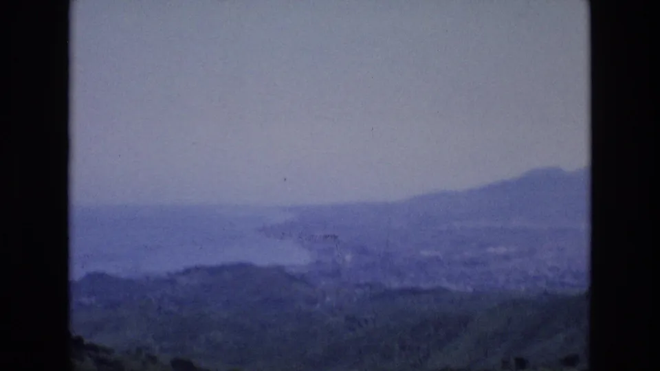 1972 Spain home-movie frame with a soft blue coastal landscape, distant ridges, water, and an indistinct bright speck or reflection near the middle distance.