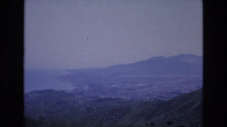 Blue-tinted 1972 Spain 8mm frame looking from a hillside toward a broad hazy bay, with distant landforms and faint town-like shapes along the shore.