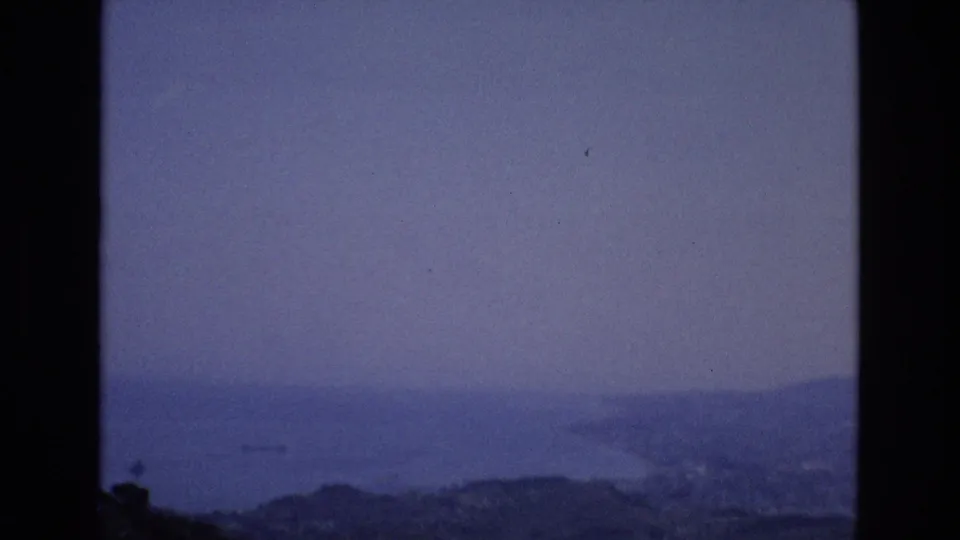 1972 Spain home-movie frame showing a muted coastal panorama with water and distant hills, plus a small indistinct dark shape near the left side of the water.