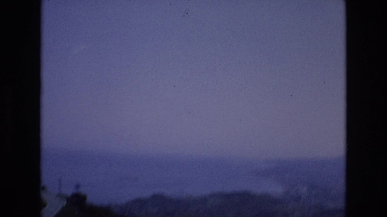 Spain 1972 8mm frame showing a hazy ocean horizon, distant shoreline, and dark foreground ridge under purple-blue dusk light.