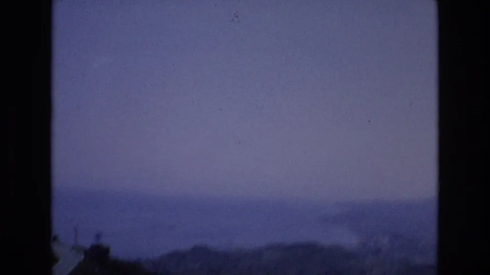 Spain 1972 8mm frame showing a hazy ocean horizon, distant shoreline, and dark foreground ridge under purple-blue dusk light.