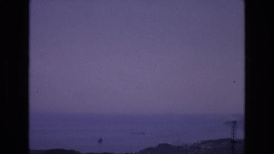 Spain 1972 8mm frame showing a dim ocean horizon, faint small boats, low shoreline, and a possible coastal road or structure in the lower distance.