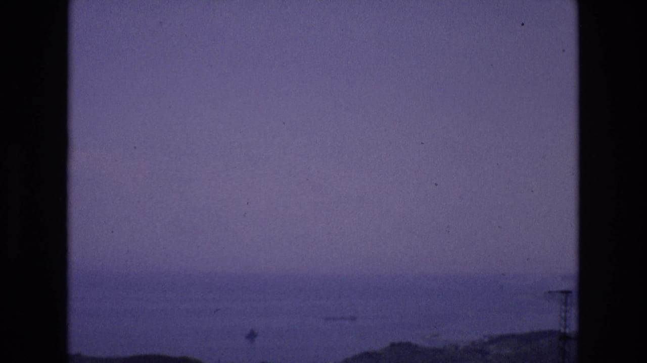 1972 Spain 8mm frame showing a distant calm sea, hazy purple-blue dusk sky, low shoreline, and dark scanned film edges.