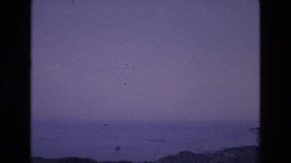 1972 Spain 8mm frame with a hazy coastal horizon, calm blue water, faint distant boat-like shape, and visible film gate edge.