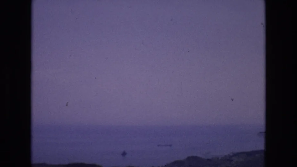 1972 Spain 8mm frame showing still coastal water, a low dark shoreline, muted purple sky, and visible dust and grain from the film scan.