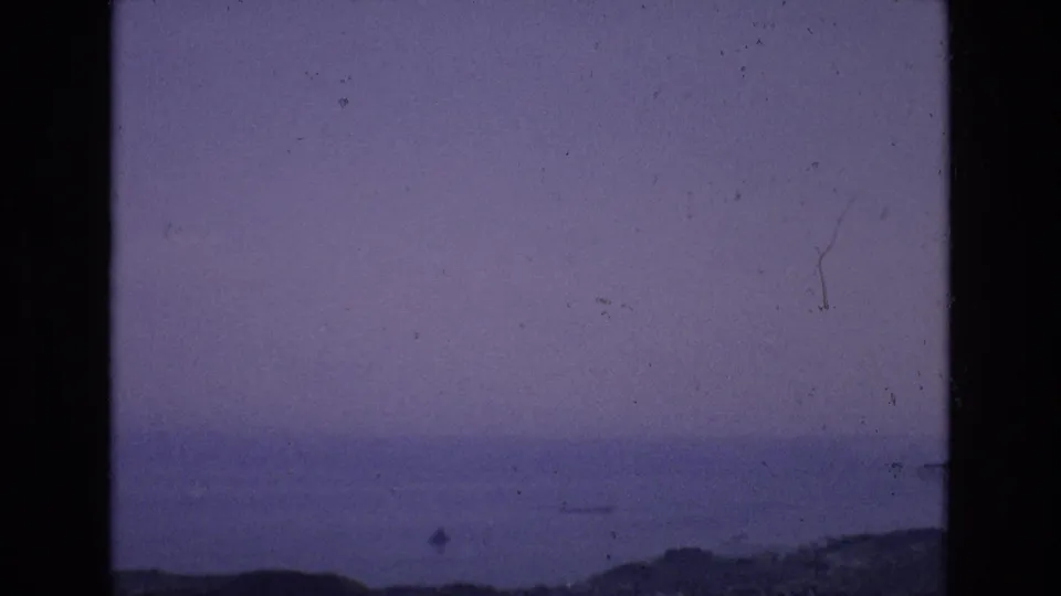 1972 Spain 8mm frame showing a quiet sea at dusk, distant low land, faint boat-like forms on the water, and blue-purple color fading.