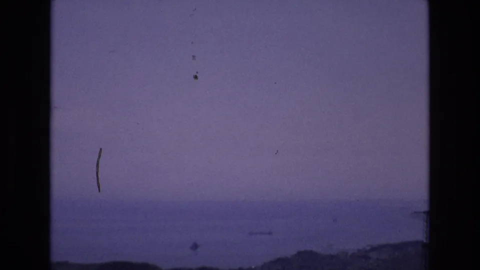 Spain 1972 8mm home-movie frame with a faint water horizon, low dark landform, blue-purple color cast, and visible dust and scratch marks.