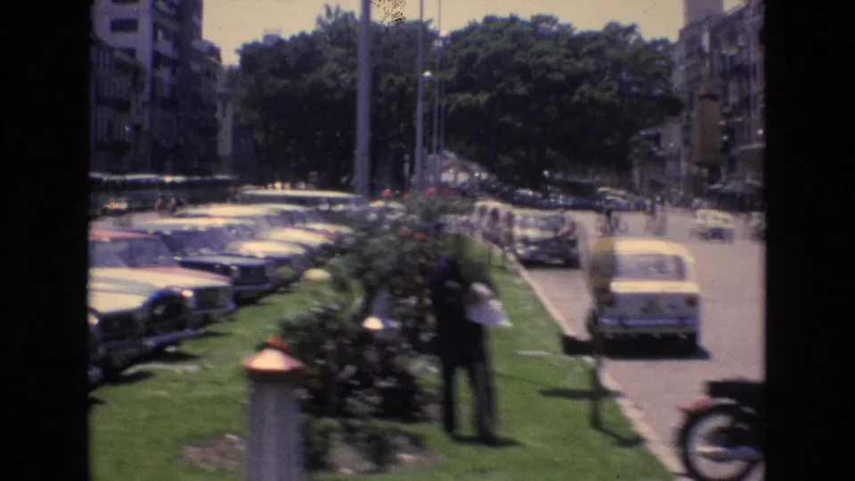 Spain 1972 street view with older cars parked beside a grassy median, a uniformed traffic officer near a planted bed, trees, surrounding buildings, and traffic on the road.