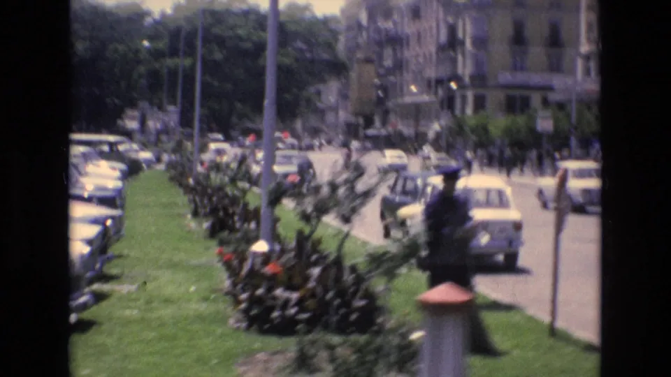 Early 1970s Spanish city street with parked cars along a grassy planted median, pedestrians in the distance, and a uniformed traffic officer near traffic.