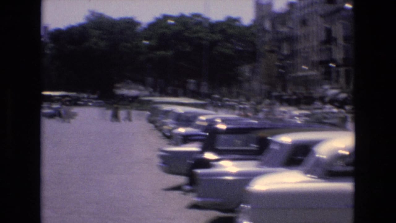 Sunlit Spanish urban parking or roadway view with rows of 1970s cars, shaded trees, buildings, and blurred vehicles passing in the foreground.
