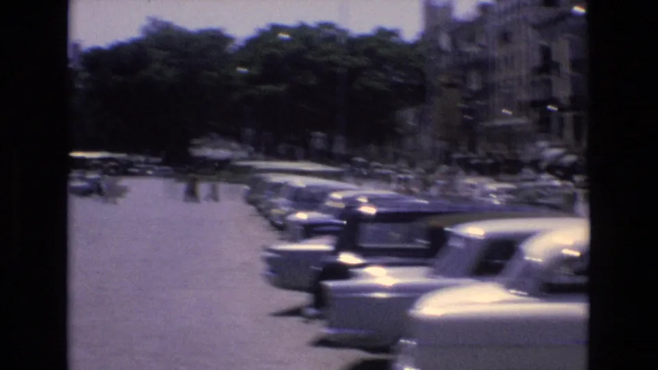 Sunlit Spanish urban parking or roadway view with rows of 1970s cars, shaded trees, buildings, and blurred vehicles passing in the foreground.