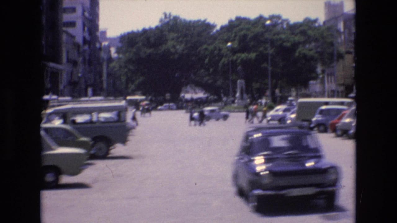 Urban roadway with parked cars, a dark car driving through, trees, buildings, and distant pedestrians.