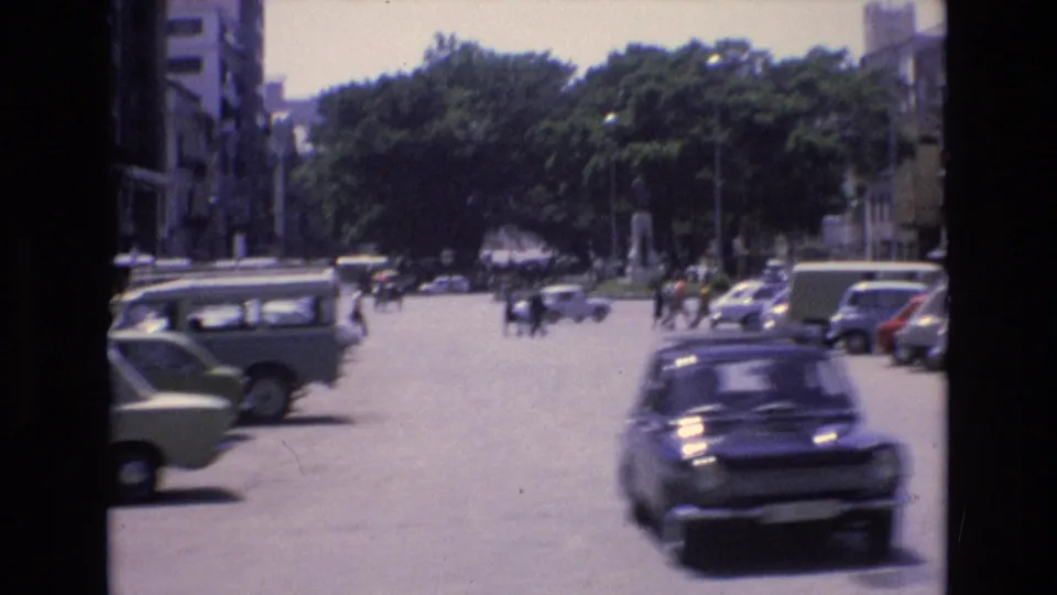 1972 Spain home-movie view of a city street with parked vehicles on both sides, a dark compact car in motion, trees, buildings, and distant pedestrians.