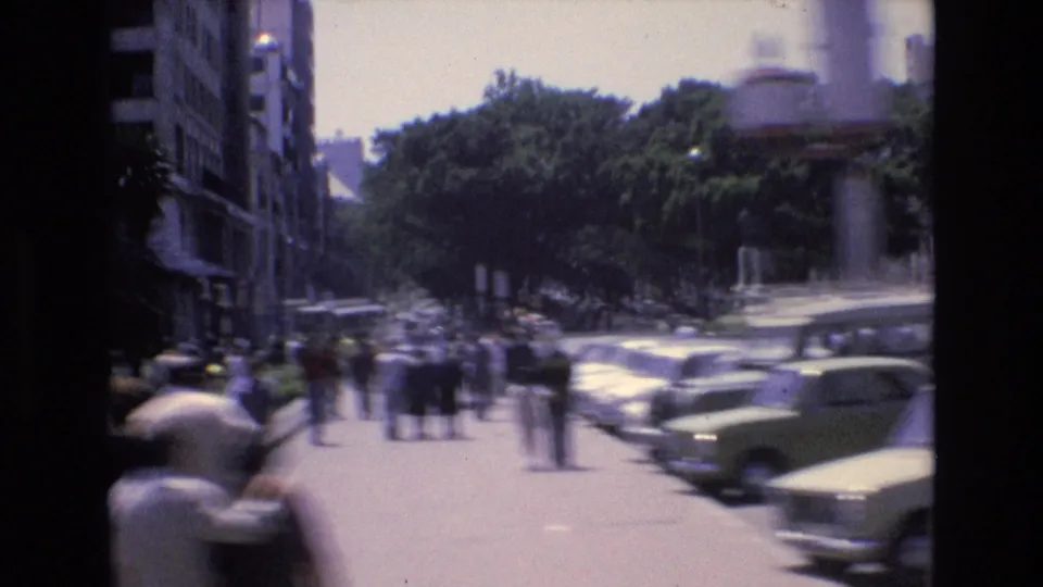 Spanish city street in bright daylight with parked 1970s cars, pedestrians in the roadway and on sidewalks, trees, buildings, and handheld motion blur.
