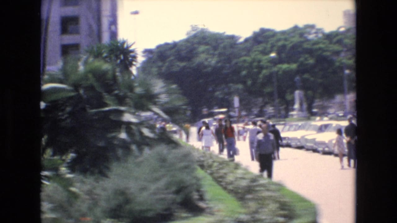 In Spain archival 8mm footage from 1972, pedestrians move along a bright sidewalk lined with trees, shrubs, palms, and parked cars near urban buildings.