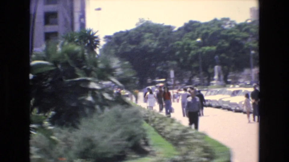 In Spain archival 8mm footage from 1972, pedestrians move along a bright sidewalk lined with trees, shrubs, palms, and parked cars near urban buildings.