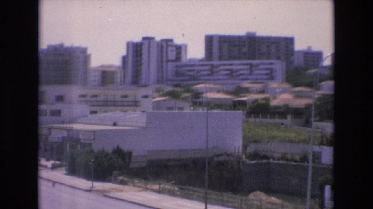 Urban buildings and low walls overlook a road, greenery, rooftops, and streetlights.