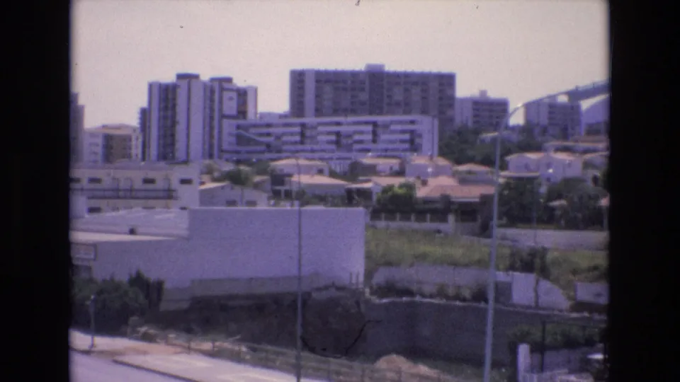 The frame holds on a Spanish urban district in 1972, with apartment-like blocks, low walls, small rooftops, a grassy area, and streetlights along a road.