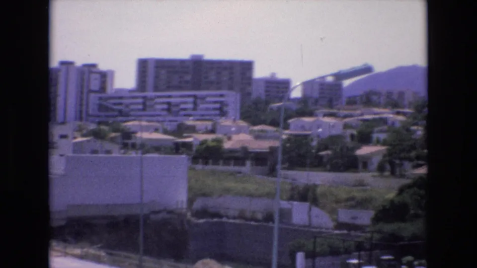 A soft 8mm view of 1972 Spain shows modernist buildings and rooftops with greenery and streetlights, plus a distant curved structure near the horizon.