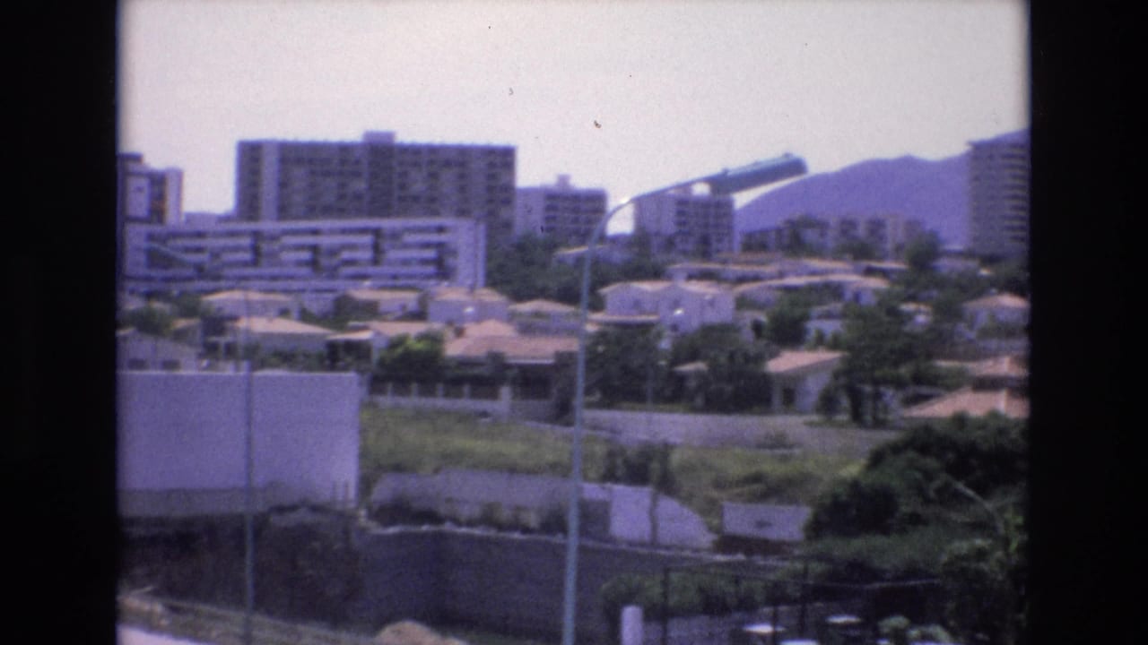 1972 Spain 8mm frame showing a sunlit urban district with mid-rise apartment blocks, low houses, greenery, foreground lamp poles, and hazy hills beyond.