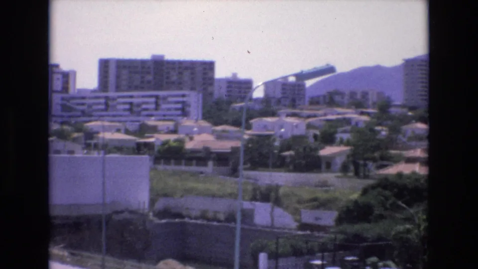 1972 Spain 8mm frame showing a sunlit urban district with mid-rise apartment blocks, low houses, greenery, foreground lamp poles, and hazy hills beyond.