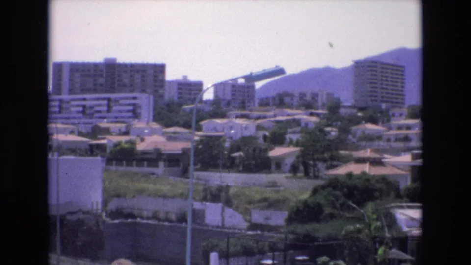 1972 Spain 8mm frame of apartment blocks and tiled-roof houses set among trees, with foreground street lighting and hazy mountains in the background.