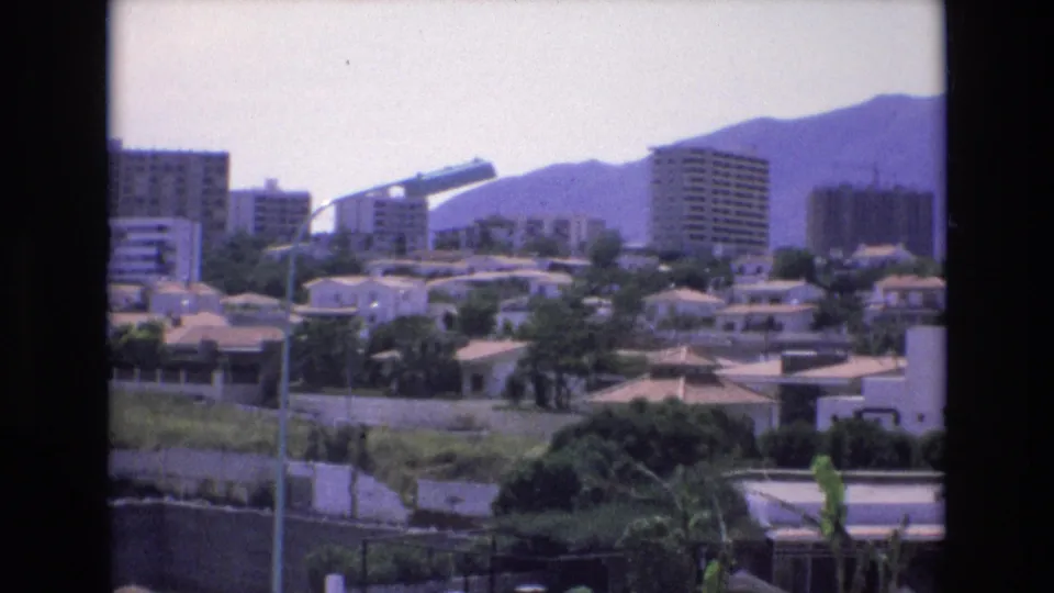 1972 Spain 8mm frame showing a residential urban panorama with tiled-roof houses, several apartment towers, green vegetation, foreground walls, and mountain slopes behind.