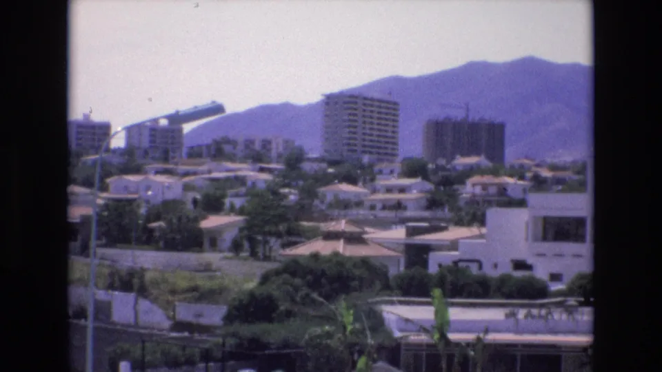 1972 Spain 8mm frame of a sunlit residential cityscape with mid-rise apartments, low tiled houses, greenery, foreground walls and lamps, and hazy hills in the distance.