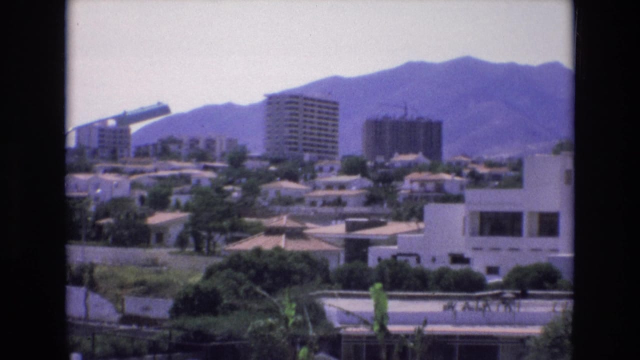 Spain 1972 home-movie frame showing an elevated cityscape with apartment towers, low residential buildings, greenery, and purple-toned mountains behind them.
