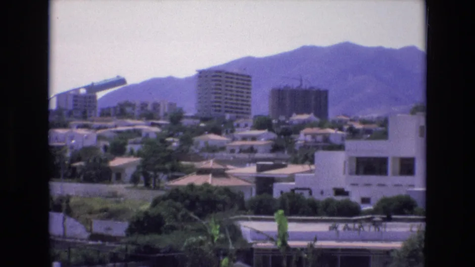 Spain 1972 home-movie frame showing an elevated cityscape with apartment towers, low residential buildings, greenery, and purple-toned mountains behind them.