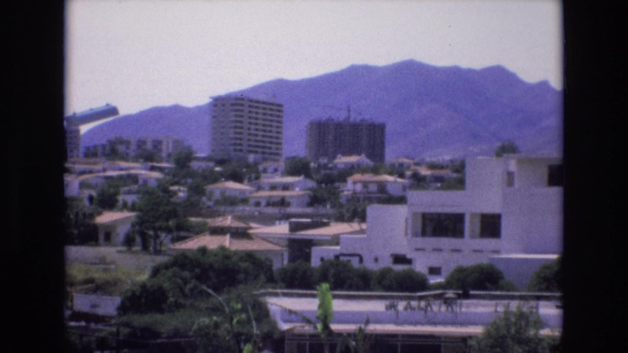 Urban buildings and trees seen below a mountain range under bright sky.
