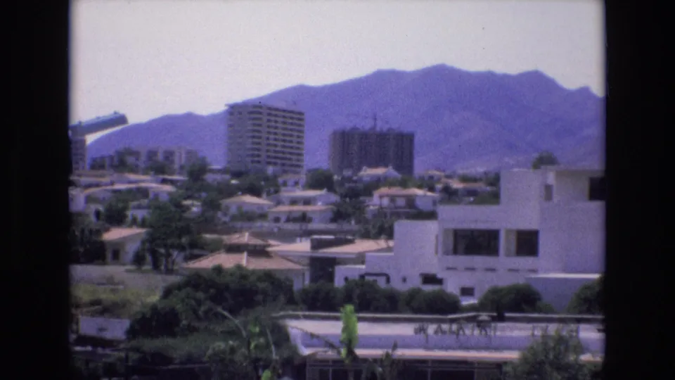 Spain 1972 frame with a steady home-movie view of high-rise residential blocks, white houses, trees, and distant mountains.