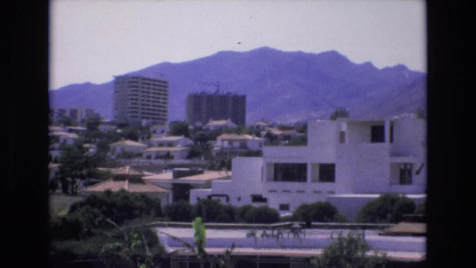 Spain 1972 home-movie still showing a white modernist foreground building, distant apartment towers, trees, and a hazy mountain ridge.